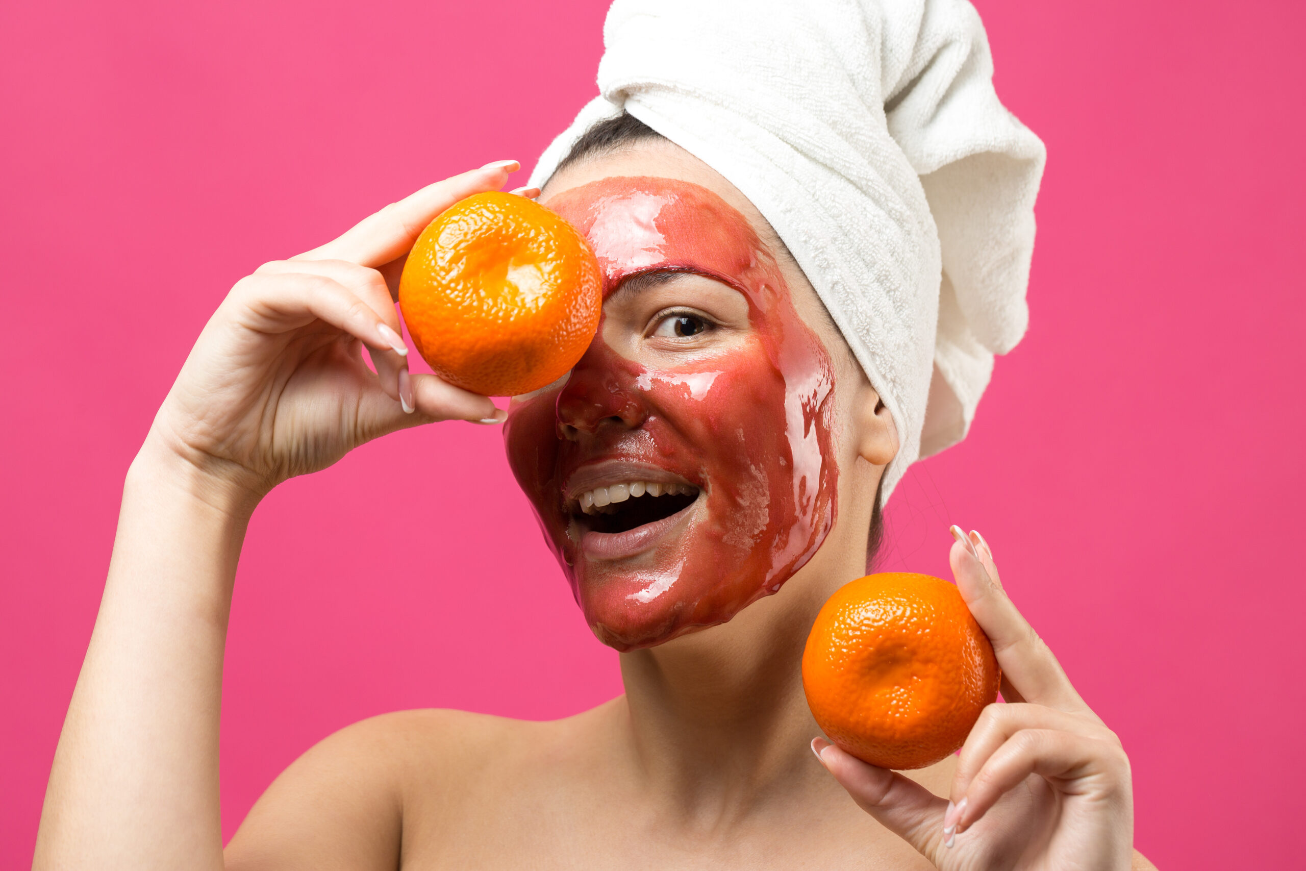 Beauty portrait of woman in white towel on head with red nourishing mask on face. Skincare cleansing eco organic cosmetic spa relax concept. A girl stands with her back holding an orange mandarin.